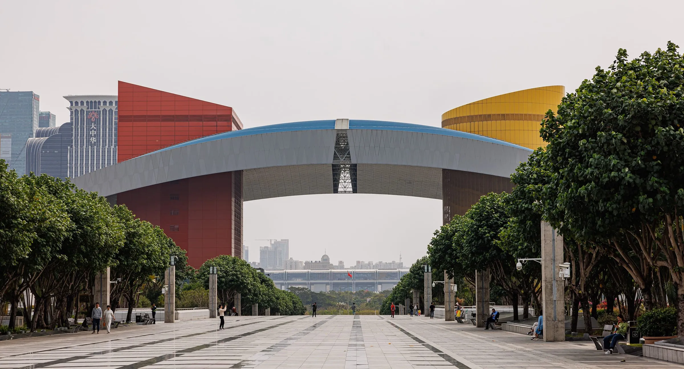 A long colonnade arch over a paved plaza in Shenzhen, with red and gold buildings behind trees
